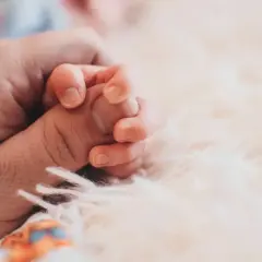 Dłoń dorosłego trzyma dłoń niemowlęcia na miękkim tle.